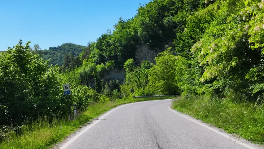 A first-person perspective drive follows a winding asphalt road through lush Italian green hills