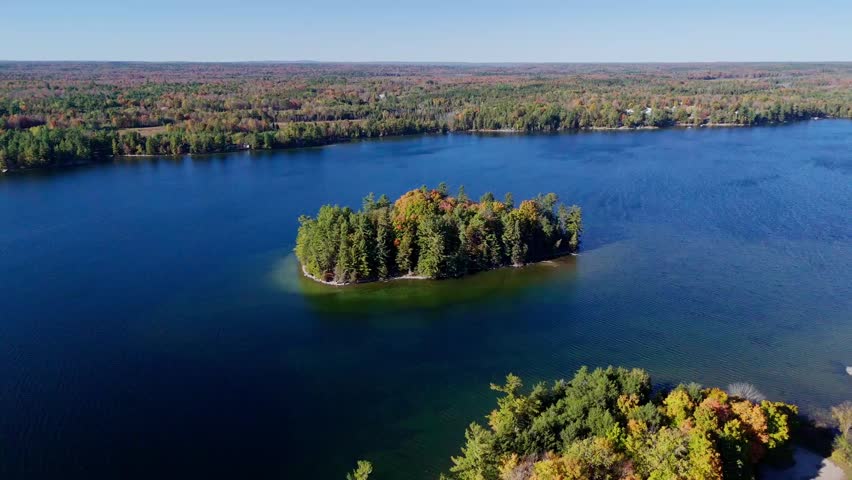 Cinematic drone shot flying sideways past a small isolated island with colorful autumn foliage in Ontario. Visible wind ripples on the lake and beautiful fall colors in the wilderness.