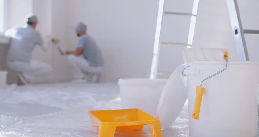 Large bucket of paint, along with small tray and paint roller, stands in foreground, ready for use. In background, two male repairmen are painting walls. Renovation and renovating house concept.