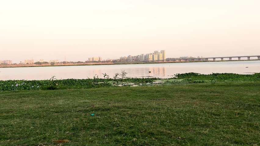 Panoramic View of Serene River Bank with Green Lawn and Distant City Skyline and Bridge at Sunset