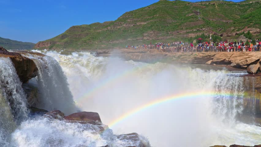 Cinematic wide-angle view of the Hukou Waterfall on the Yellow River. Powerful water cascades into the gorge, creating a prominent double rainbow against a backdrop of rocky cliffs and tourists.