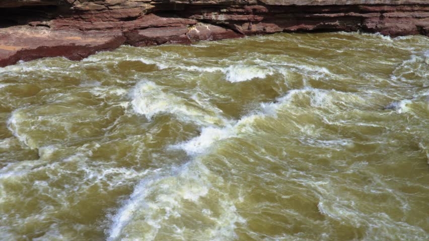 Detailed pan from the churning, silt-heavy rapids of the Yellow River to the deep rocky gorge of Hukou Waterfall. Features the red sandstone cliffs and rushing white water.