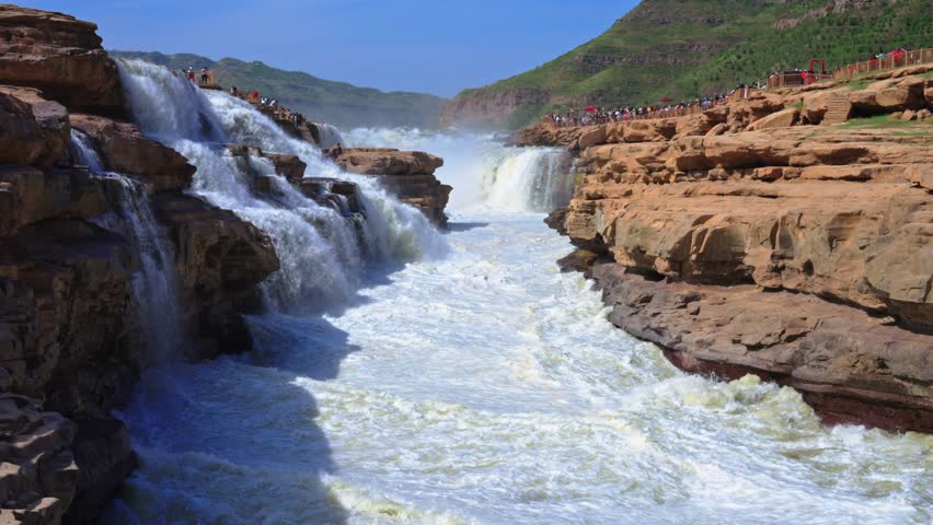 Dynamic panning shot of the Yellow River rapids flowing through the red sandstone gorge at Hukou. Features the turbulent white water and layered sedimentary rock.
