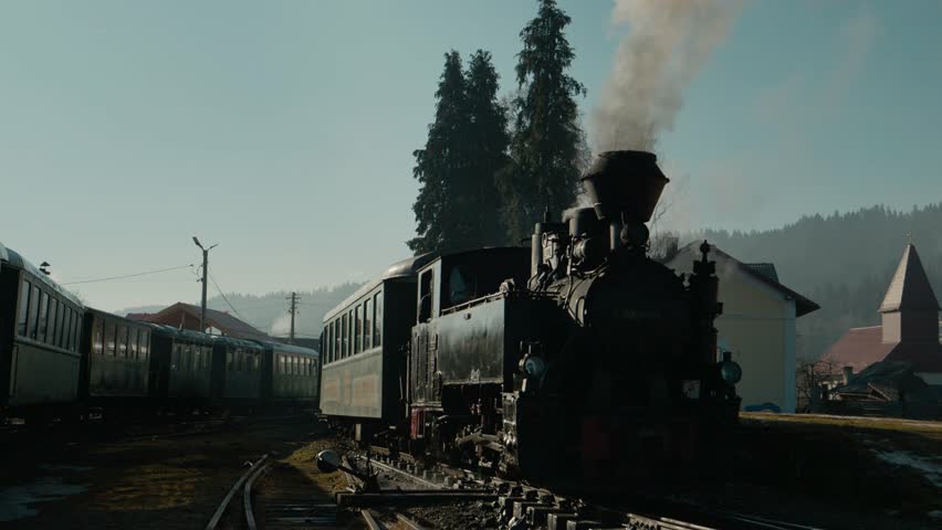 The Mocanita steam train stands at the station in Bucovina, Romania. Smoke rises from the chimney as the sun shines on the old train and surrounding area.