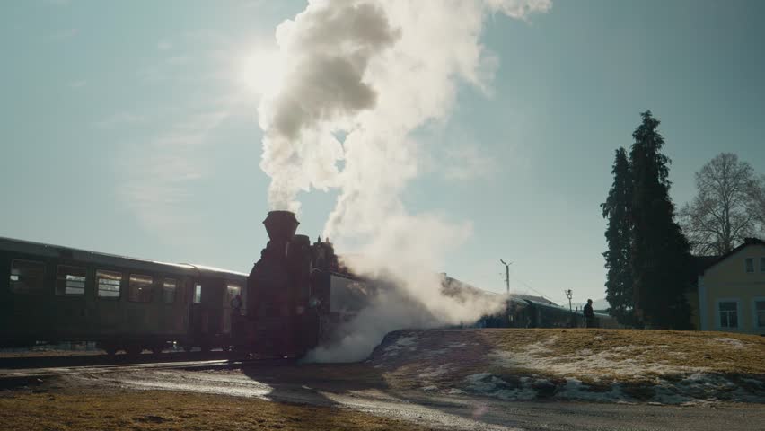 Mocanita steam train is parked at Bucovina station in Romania. Smoke rises from the train as the sun shines brightly in the sky. People are nearby observing the train.