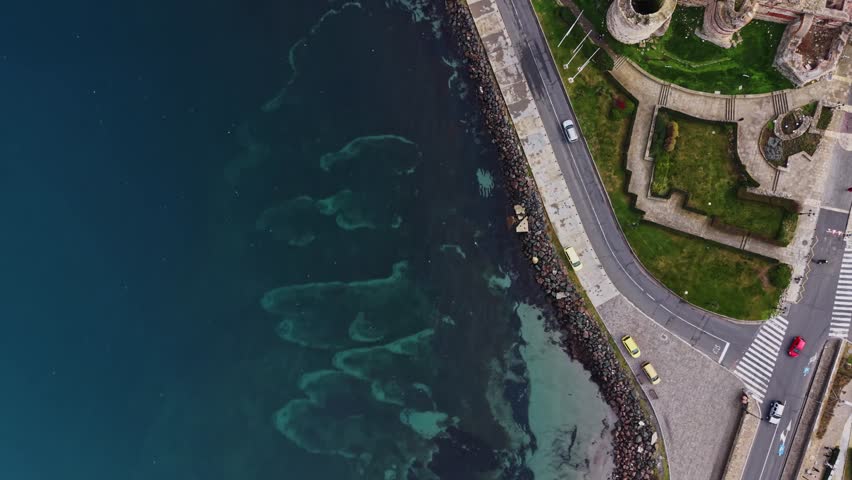 Nesebar showcases its waterfront with ancient ruins nearby. Boats are docked by the shore, and people are seen along the pathway enjoying the sunny weather and ocean view.