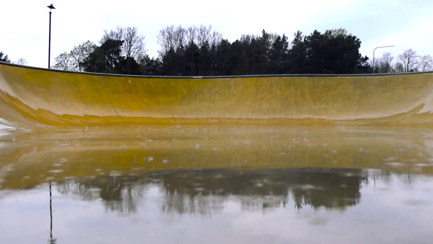 Slow motion view of raindrops falling into a large puddle on a yellow skatepark ramp, creating ripples on the water surface with reflections of trees and an overcast sky