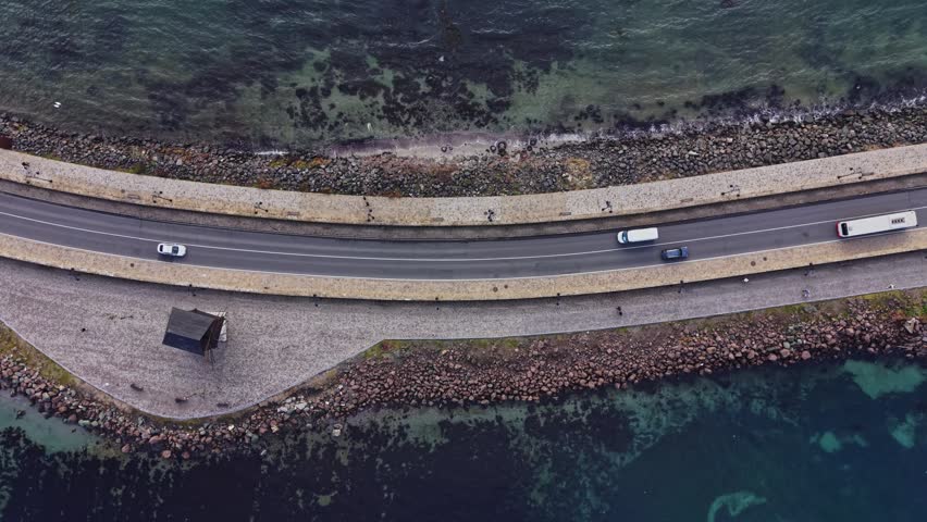 Cars travel along a coastal road in Nesebar, Bulgaria. The asphalt path runs parallel to the sea, showing the shoreline and a small building nearby.