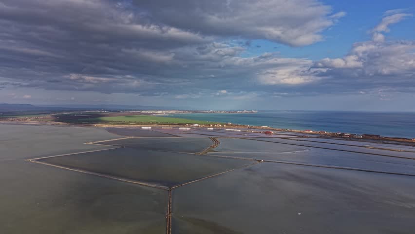 Workers are seen in the salt mining area by the coast. Large salt flats are visible in the foreground with water pools reflecting the sky.