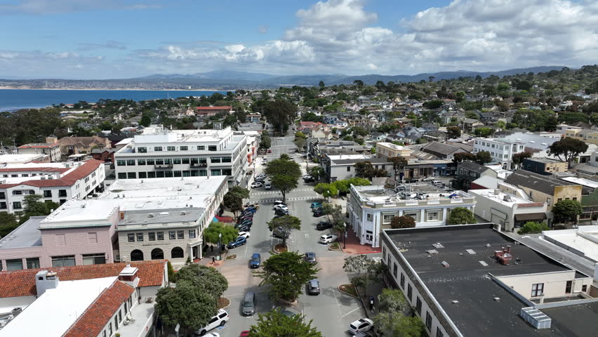 Cinematic aerial drone footage moving sideways above Lighthouse Avenue in Pacific Grove, California, showcasing coastal neighborhoods, downtown buildings, oceanfront streets, Lovers Point Park in the
