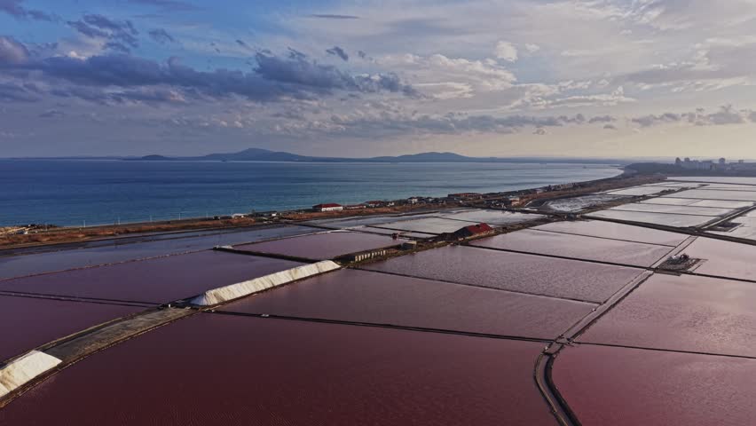 Workers are seen in the salt mining area by the ocean. Large salt pans stretch across the landscape, reflecting the sky and surrounded by distant mountains and buildings.