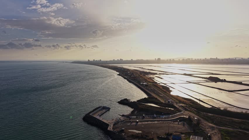 The scene shows a vast coastline with water and land meeting in the late afternoon. The sun shines over the horizon, illuminating the shoreline and nearby city structures.