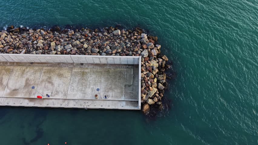 A concrete pier extends into the water with rocks along the edge. Some red buoys float nearby. The scene is captured from above during daylight hours with clear skies.