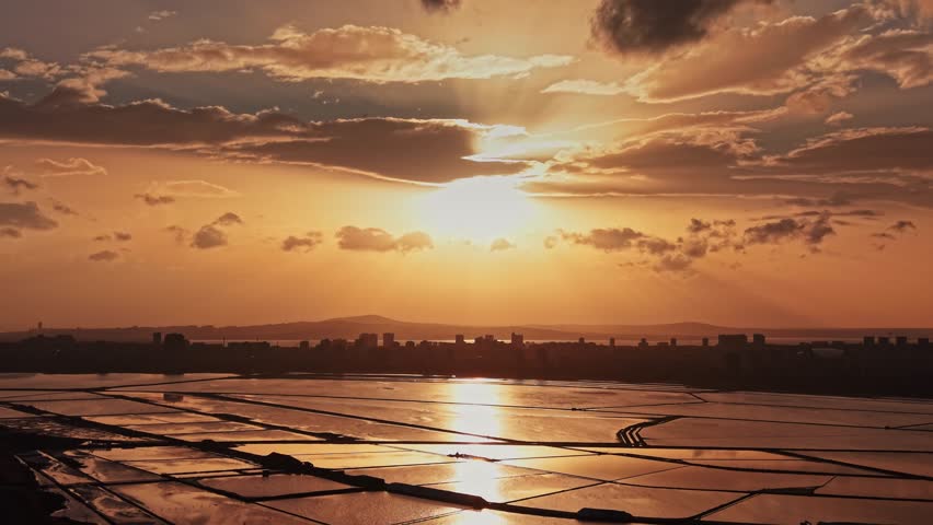 The sun sets over a landscape of water and land. Clouds fill the sky and reflect light over the water. Buildings are visible far in the background.