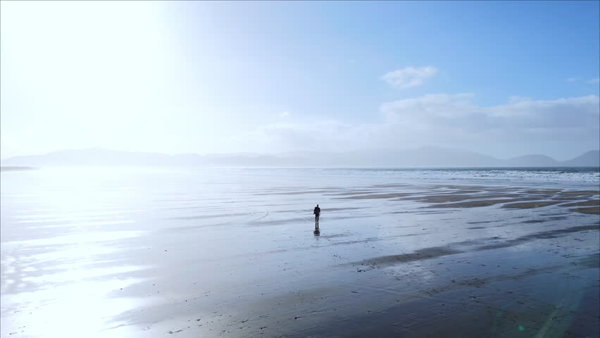 A static aerial shot at Inch Beach in County Kerry, Ireland, showing a person walking across shallow reflective water on the sand with blue sky, clouds, mountains and the sea in the background.