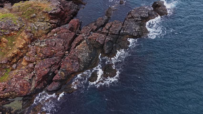 Waves crash against rugged rocks along the coastline. The shore features a mix of dark stone and vibrant colors. Water flows gently around the rocky formations under bright skies.