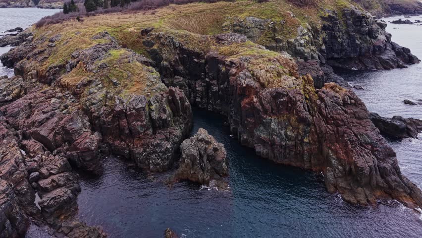 The scene shows a rocky coastline with water flowing around the land and patches of grass growing on the rocks in a natural setting. The sky is overcast and the location is near the shore.