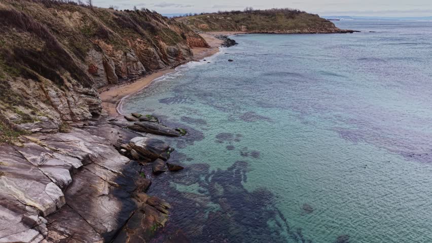 A coastal area with clear water near a sandy beach. Smooth rocks are visible along the shore, and hills lie in the background under a cloudy sky.