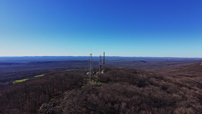 Communication towers stand on a mountain surrounded by trees. The sky is sunny with few clouds. The landscape extends far into the distance, showing hills and valleys.
