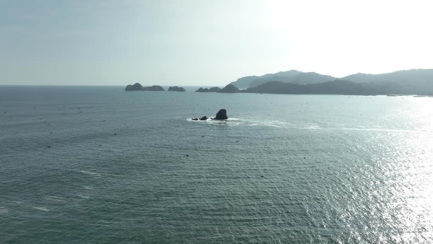 Distant rock island in calm sea with sunlight reflecting, mountains on the horizon