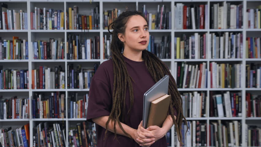 Positive young woman with dreadlocks smiling happily while holding a laptop and a book in a library