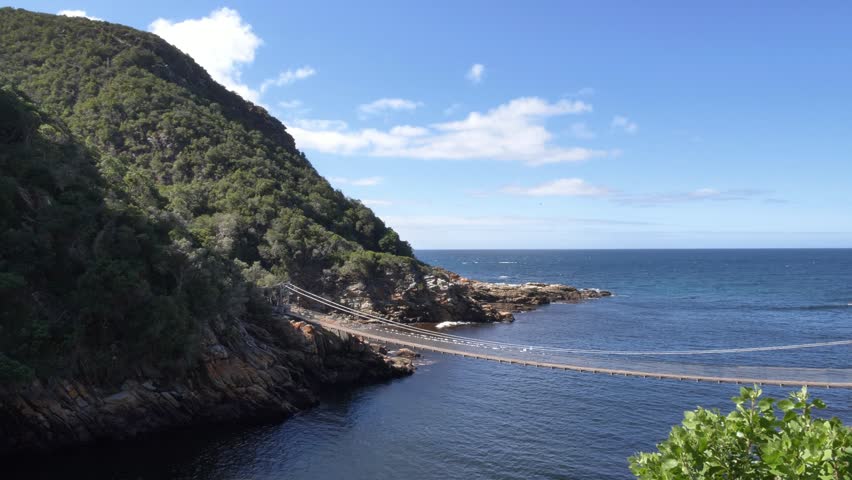 A scenic view of a suspension bridge connecting two rocky shores, with the deep blue ocean stretching to the horizon under a clear sky at Storm’s River Mouth, Tsitsikamma National Park, South Africa.