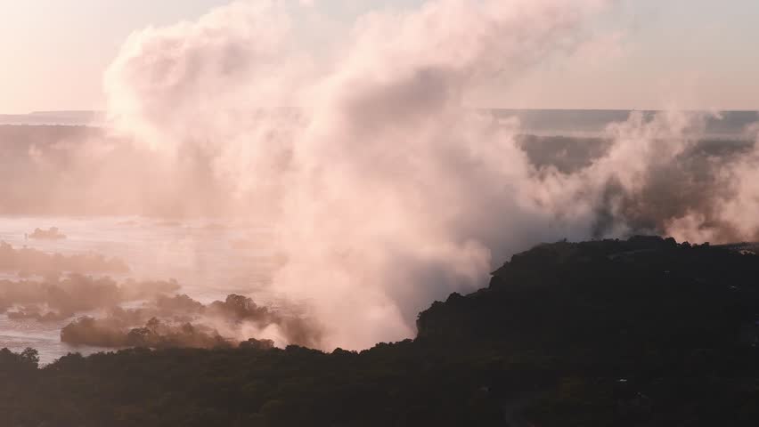 Aerial sunset view of Victoria Falls in Zimbabwe shows the Zambezi River, a rising spray plume that drifts and shifts, shadowed cliffs, islets, and golden hour light.