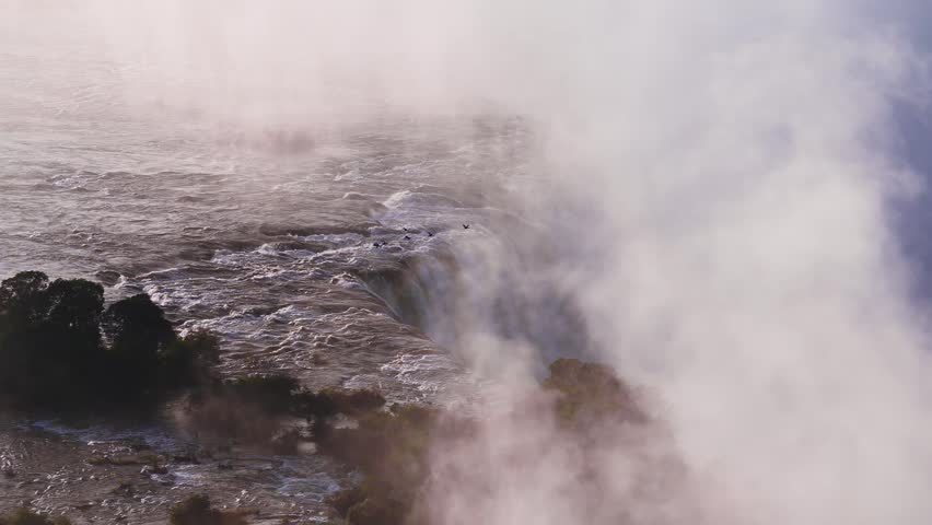 Aerial view shows Victoria Falls as water tumbles over the lip, mist rises, rapids churn, and birds arc through vapor near tree covered islands at sunset.
