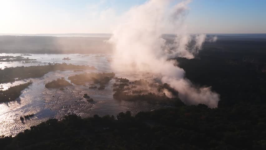 Aerial view passes Victoria Falls in Zimbabwe as mist plumes rise, braided channels flow, islands dot the Zambezi, and warm late afternoon light reflects on water.