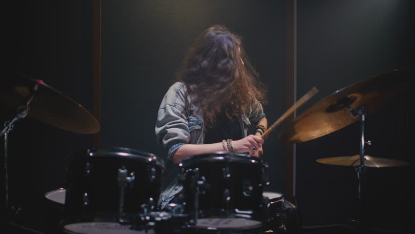 Young female drummer passionately playing a drum kit in a professional rehearsal studio, energetically striking cymbals and drums with drumsticks. Hair flying. Rock Feminist style. Woman power.