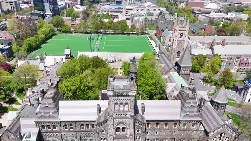 Aerial view flying backward, pulling away from University College to reveal its full stone facade, surrounding campus buildings, and lawn
