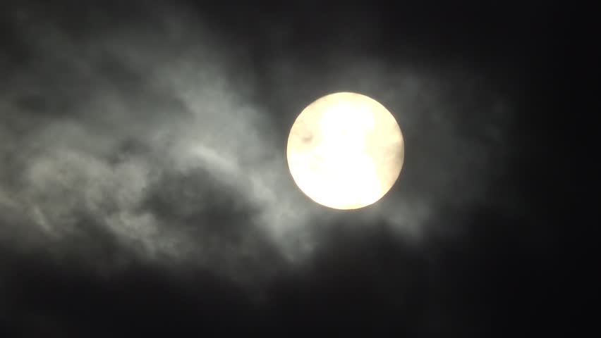 Close up shot of the bright afternoon sun appearing as a clear glowing disk while thin dark wispy clouds drift across it.