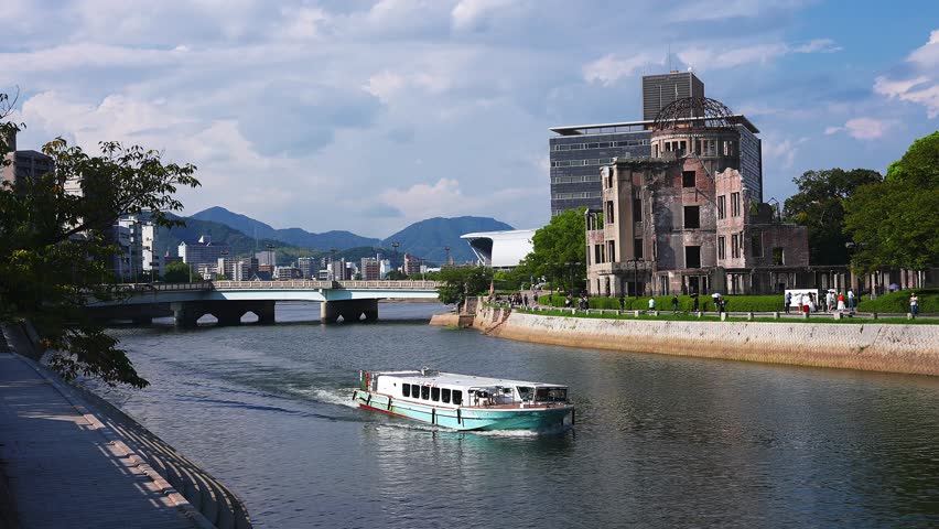 Midday view in Hiroshima, Japan, shows the Atomic Bomb Dome, a low arched bridge, a sightseeing boat on the Motoyasu River, pedestrians, offices, and parkland.