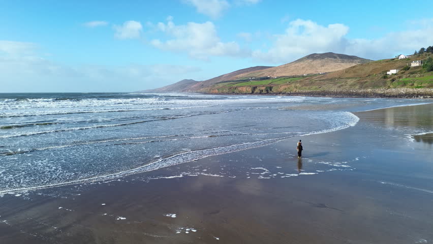 A circular aerial shot orbiting around a person riding a horse on Inch Beach in County Kerry, Ireland, with reflective wet sand, blue sky, clouds and mountains creating a scenic coastal backdrop.