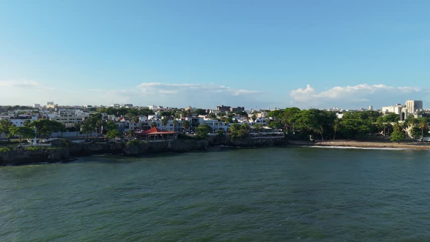 Lateral aerial view of the coastal city skyline, rocky shore, and Caribbean Sea waves during a bright day in Santo Domingo, Dominican Republic