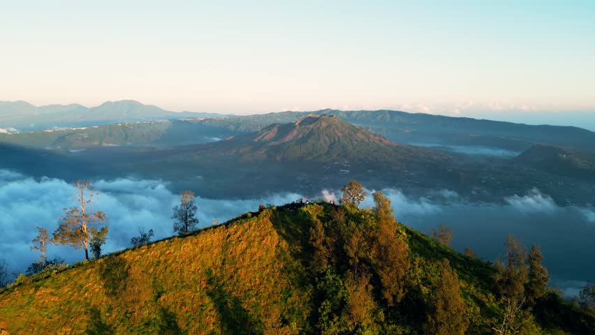 Volcanic mountain terrain with crater lake and grassy ridge in golden morning light with low clouds and long tree shadows across remote highland landscape smooth descending aerial drone flight