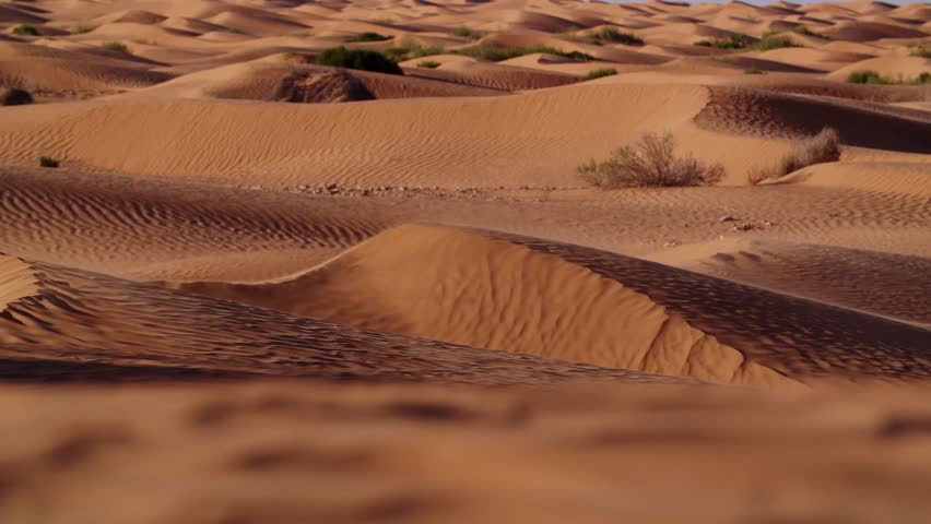 Cinematic drone footage of the Sahara Desert in Tunisia at sunset, with strong winds blowing sand across the dunes