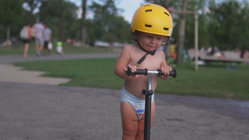 Toddler riding scooter in park wearing yellow helmet, little child exploring paved path with playful curiosity, enjoying sunny outdoor summer afternoon
