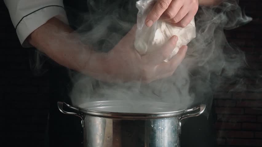 Chef hands in a white uniform lift a plastic bag filled with frozen dumplings, pouring them into a steaming pot of water, with vapor rising in a rustic kitchen environment.