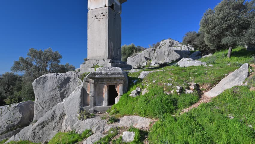 Ancient Lycian pillar tomb standing amongst rocky ruins and green grass under a clear blue sky in the historic city of Xanthos, Turkey