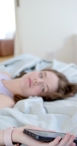 Vertical video: Woman lying on bed in purple tank lifting black phone to right ear answering call. Smartphone, smartwatch, bedroom, blanket, pillow, lifestyle, vertical video