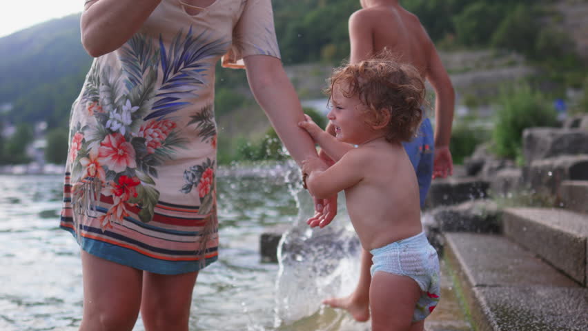 Toddler standing by lake reaching toward mother’s bracelet while older sibling stands nearby, showing curiosity, interaction, and family bonding in a summer outdoor setting