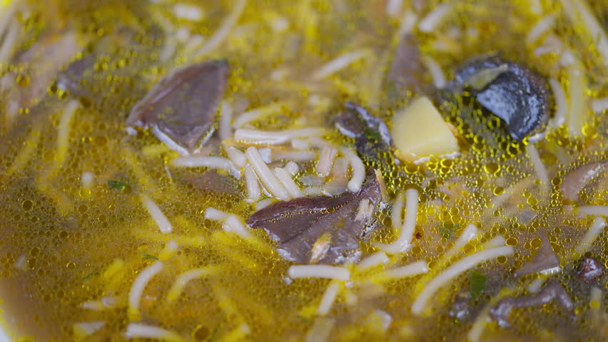 Noodle soup simmering. Aromatic mushroom noodles in warm flavorful broth. Closeup of bubbling mushroom noodle soup with aromatic spices and golden broth in slow motion