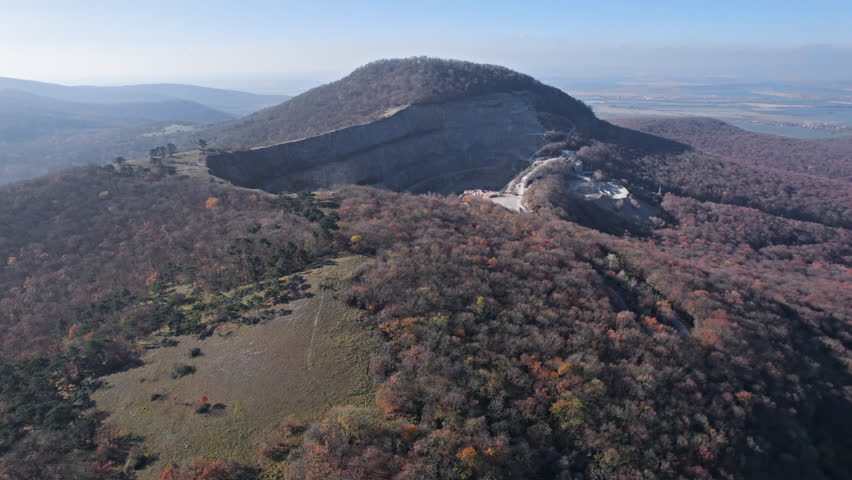 Flight over of a large open pit quarry with terraced excavation roads surrounded by forested hills in a rugged autumn landscape, Slovakia.