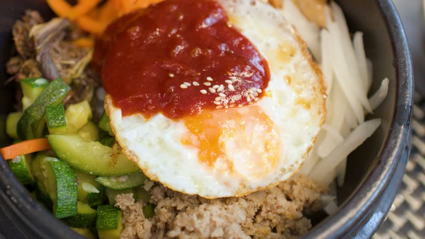 Close-up shot of a spoon mixing rice, vegetables, and egg yolk in a stone bowl