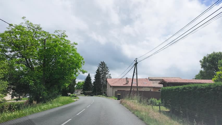 A vehicle travels along a paved road past stone houses and green trees under clouds