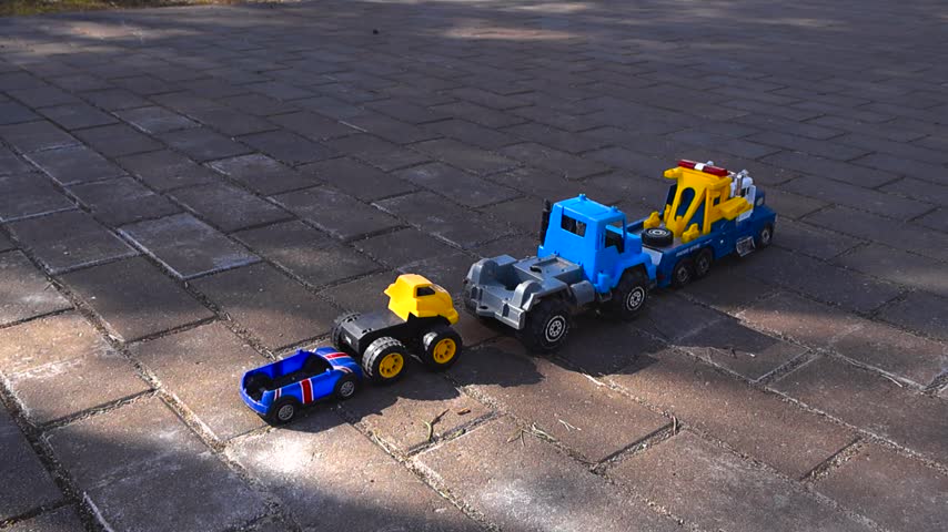Colorful toy trucks and a race car forming a convoy on a paved ground outdoors during a sunny day. A blue carrier is towing another truck, showcasing a childhood play scene with vehicles