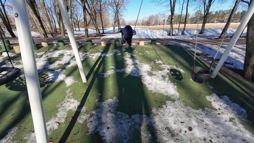 A child enjoys swinging in a snowy winter park, surrounded by bare trees. The clear blue sky creates a bright, cheerful atmosphere. Captured in crisp daylight with vibrant colors.