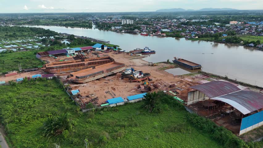 An aerial view of a shipyard in operation, featuring large rusting ships and barges under construction on dry land along the banks of the Kapuas River in West Borneo, Indonesia.