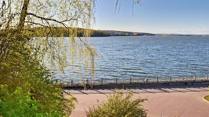 Scenic lakeside walkway lined with trees and bushes, overlooking calm water and distant forested hills beneath a clear blue sky, creating a tranquil natural landscape